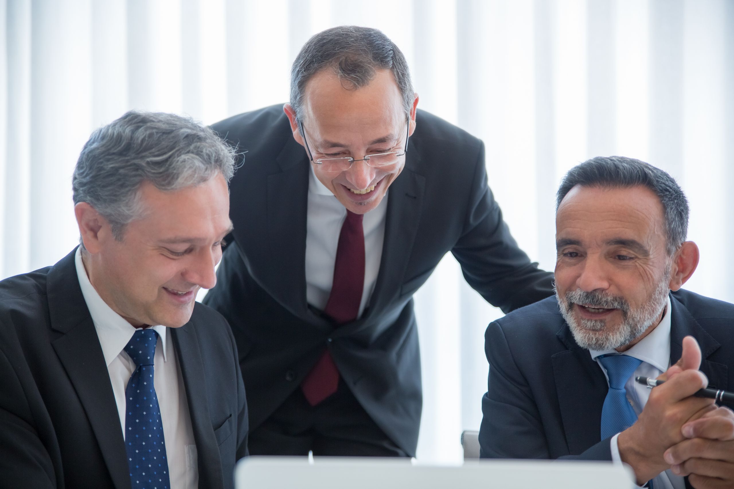 Home Three businessmen in suits engaging in teamwork indoors with smiles.